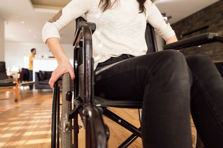Unrecognizable young disabled woman in wheelchair at the at home in her living room. Close up of her arm laid on wheel.