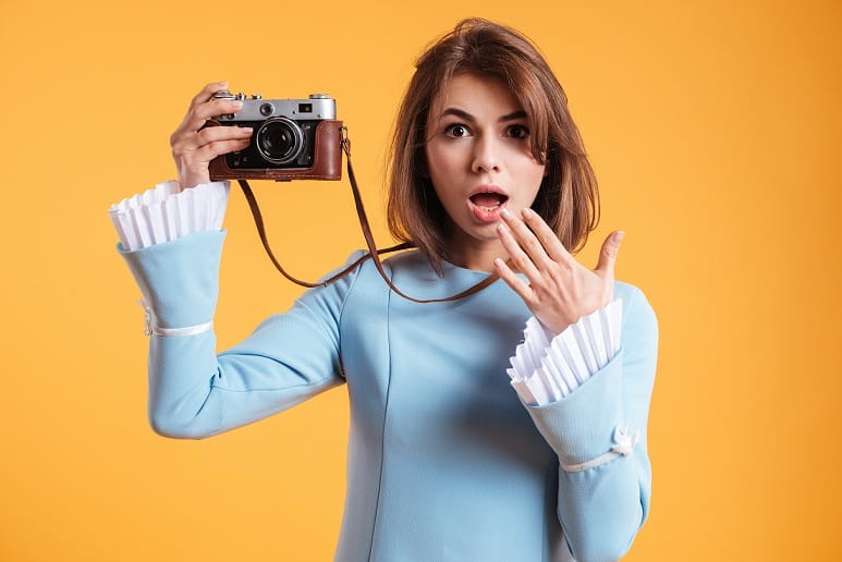 Amazed young woman taking pictures with old vintage camera over yellow background